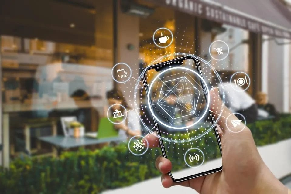 Hand holding a smartphone displaying a virtual geometric interface, surrounded by digital icons, in front of a café.