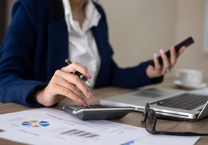 Person in a business suit uses a calculator and holds a phone at a desk with a laptop, charts, and glasses.