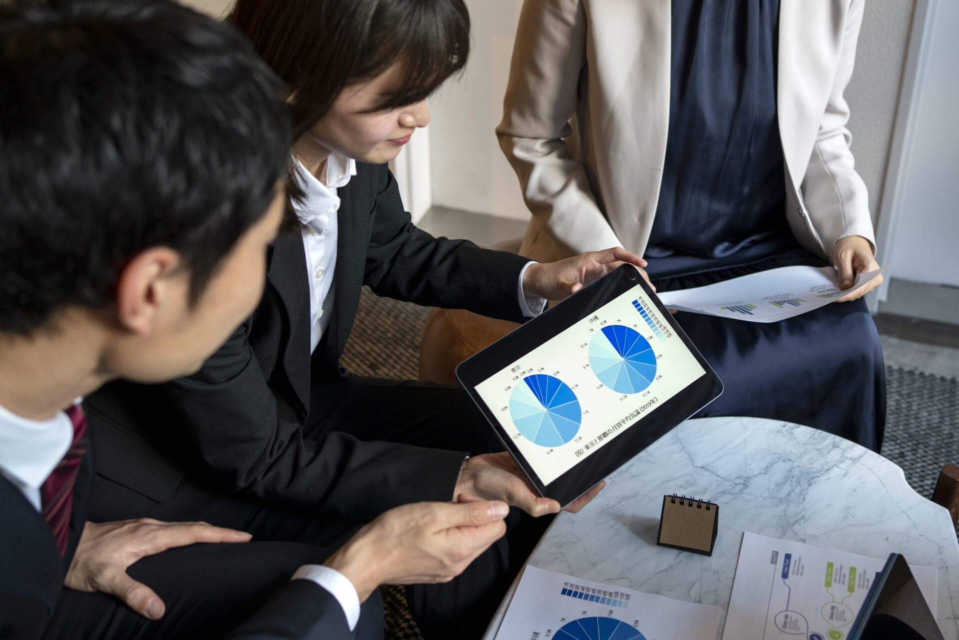 Three people in business attire discuss pie charts displayed on a tablet, focusing on database performance management strategies during a meeting at a round table.