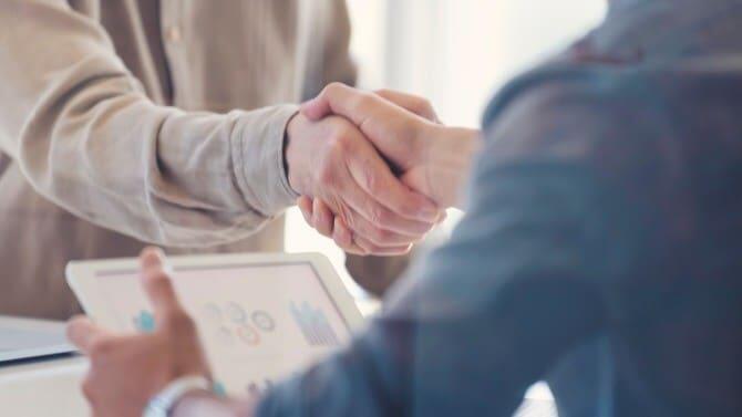 Two people shaking hands over a table, with one holding a tablet displaying graphs.