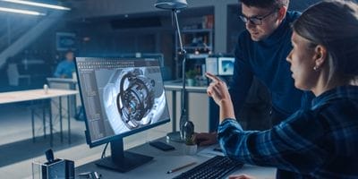 Two people discussing a 3D mechanical design on a computer monitor in an office setting.